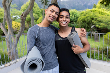 Portrait of diverse gay male couple holding yoga mats and smiling on balcony
