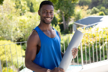 Portrait of african american man holding yoga mat smiling on sunny garden terrace