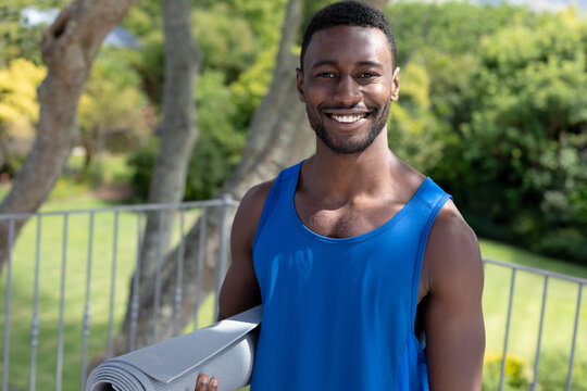 Portrait of african american man holding yoga mat smiling on sunny garden terrace