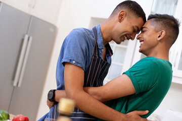 Diverse gay male couple spending time in kitchen embracing and smiling