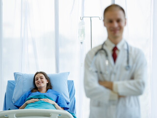 Obraz premium Focus to caucasian woman patient smiling look at camera and sleep on bed in room. Doctor man wearing uniform carrying stethoscope on neck smiling and standing folded crossed arms