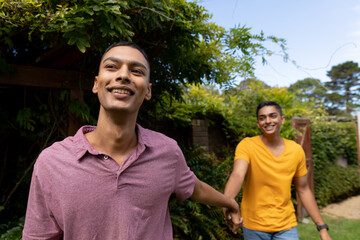 Diverse gay male couple holding hands and walking in garden