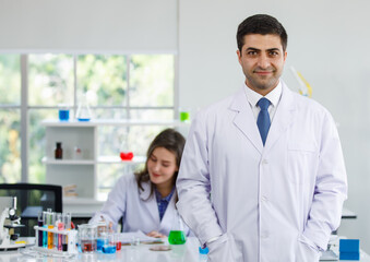 Portrait focusing on caucasian scientist man wearing uniform standing hands holding in pocket uniform and look at a camera with a smile in laboratory. Scientist woman is smiling in blurred background