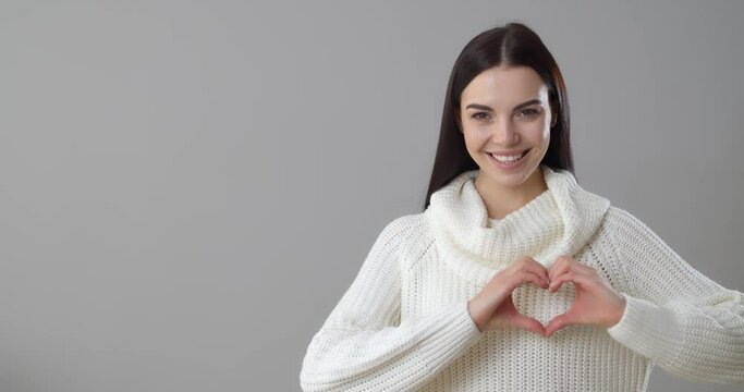 Beautiful young woman making heart with her hands on grey background