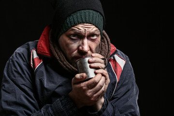 Portrait of poor homeless man with cup of hot drink on dark background