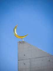 Naklejka premium Crescent moon on top of a modern mosque minaret against the vivid blue sky. A minaret is a visual focal point of a mosque and also provides vantage point to perform the call to prayer 5 times a day.