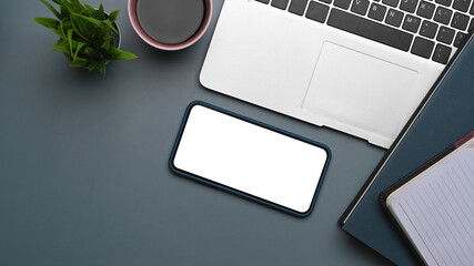 Top view of modern workspace with smart phone, laptop, coffee cup, notebook and plant on dark blue background.