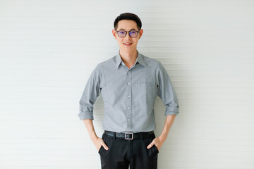Fototapeta premium Medium portrait of a formal Asian handsome man with cute smiling, wearing eyeglasses in a gray long sleeve shirt and black pants with hand in pocket posing on white background in the studio