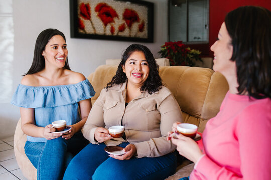 Three happy latin friends women talking and drinking coffee and tea sitting on a couch at home in Mexico city