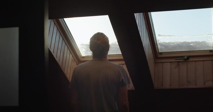 Back view silhouette of person admiring rising sun indoors slow motion. Unrecognizable man looking out of wooden window to blue sky dreaming of freedom. Loneliness solitude isolation concept 