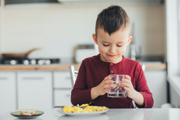 A child in the afternoon in a white light kitchen drinks water from a glass