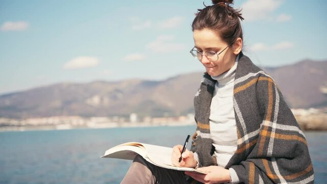 A young pensive woman writer sits wrapped in a blanket on a pier and writes notes in a notebook with inspiration.