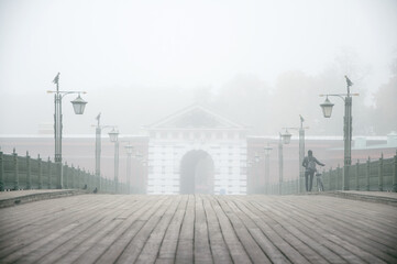 Foggy atmospheric landscape of wooden boardwalk bridge with retro elegant lanterns on the sides and silhouette of man from the back walking with bicycle to the entrance of white arch in old building