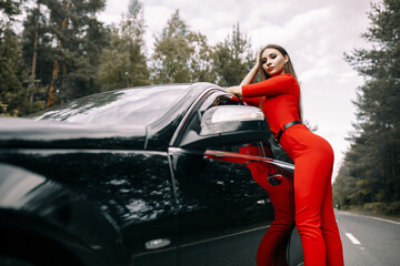 A beautiful young girl in a red overalls stands by a black car on an empty road in the forest