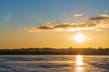 Winter landscape with frozen lake at sunrise or sunset. Lake glistening ice reflect a sun.Forest in the background.