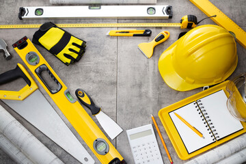 Contractor theme. Tool kit of the contractor: yellow hardhat, libella, hand saw. Plans and notebook on the gray tiles background.
