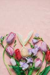 DIY Easter egg with cute face and rabbit ears framed by bouquet of colorful spring flowers: tulips and freesias, on pink background. Festive springtime flat lay. Greeting card mockup. Copy space.