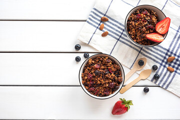 Home-made baked granola in two bowls, with berries, almonds, napkin and eco-friendly spoons. White rustic background, copy space. 