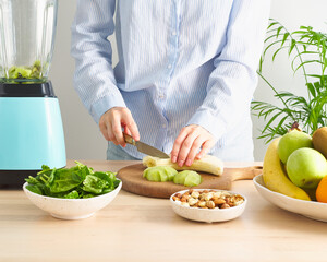 Raw foodism, green smoothie with vegetables and fruits. Woman cutting kiwi and banana to blender, dietary plant based mix for vegan or vegetarian on table in home kitchen. Low carb and gluten free