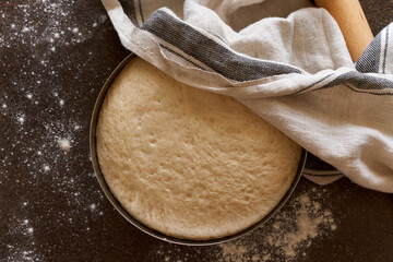 Raw yeast dough resting and rising in large metal bowl covering with linen towel on dark background. Wooden rolling pin and flour scattering on table