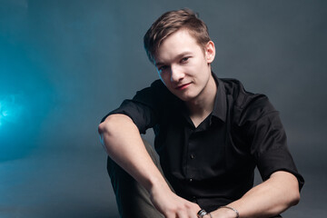 A guy with a serious face sits against a backdrop of blue smoke