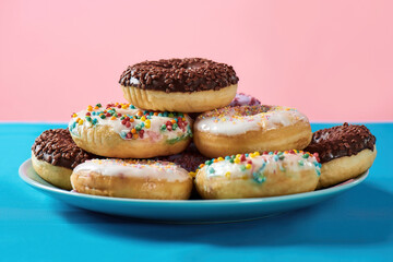 Breakfast time. Close up of various colourful round glazed donuts with sprinkles on the plate over blue and pink background