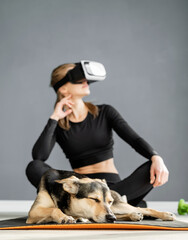 Young woman in sport clothes wearing virtual reality glasses sitting on fitness mat with dog