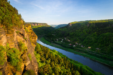 Sandstone rock formations and forests in the evening sunshine with a view of the Elbe river in the Elbe Canyon.