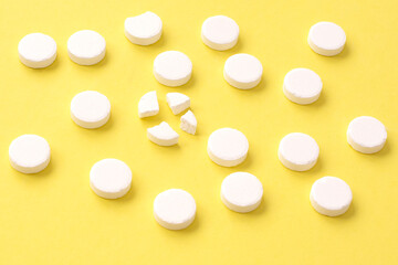 Round white medical pills arranged and one of them is crushed into four parts on yellow background....