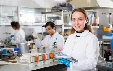 Young female scientist making research in chemical laboratory and taking notes in test chart..