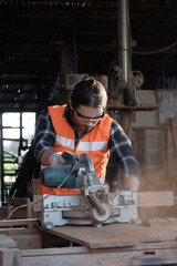 Young caucasian carpenter is working in a wooden furniture factory, cutting timber with an electric saw, dust to spread. The manufacturing industry has to be protected for professional safety.