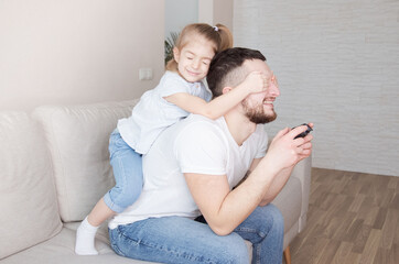 Smiling little girl covering eyes of her father while he playing computer games on sofa at home.