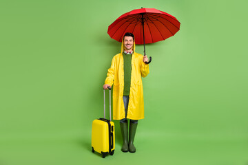 Full size photo of brunet optimistic guy with umbrella bag wear cloak boots isolated on green color background