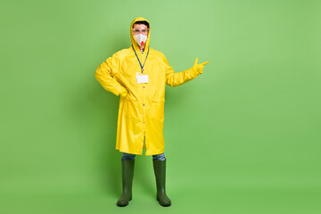 Full body photo of young man wear protective mask corona virus indicate finger empty space ad isolated over green color background