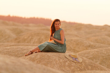 Young beautiful woman with long hair in blue dress sitting elegant and thoughtful on sand at sunrise in sandy desert