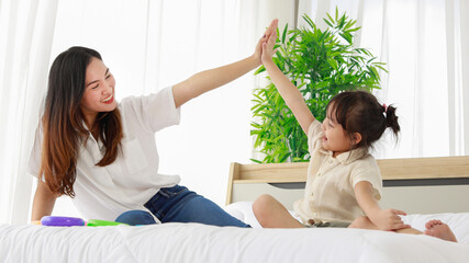 Asian black hair little playful daughter smiling touch hands hi five with beautiful young single mother sitting on white clean sheet bed with green tree pot and clear curtain behind in bedroom