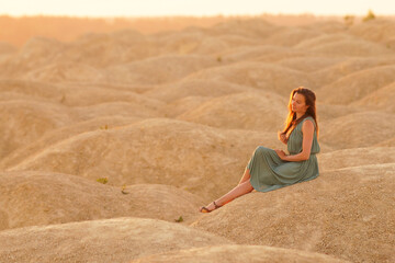Young beautiful woman with long hair in blue dress sitting elegant and thoughtful on sand at sunrise in sandy desert