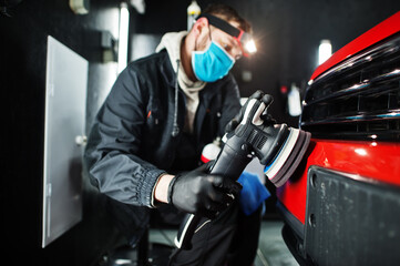 Car detailing concept. Man in face mask with orbital polisher in repair shop polishing orange suv car.