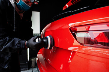 Car detailing concept. Man in face mask with orbital polisher in repair shop polishing orange suv...