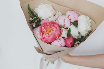 Hand holding stylish peonies bouquet in paper on background of white wall. Happy Mothers day