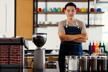 Asian short black hair young successful handsome male barista wear brown shirt and blue jeans bib apron stand smiling behind counter bar full of paper coffee cups, maker machine, beans and equipments