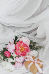 Beautiful stylish peonies bouquet and gift box on soft white fabric on rustic chair. Bridal morning