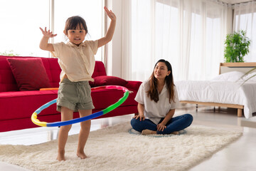 Asian beautiful young long black hair single mom stand smile playing hula hoop together with little cute daughter girl next to red sofa and bed in living bedroom with clear glass windows background © Bangkok Click Studio