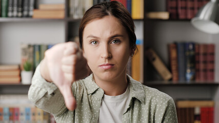 Woman thumb down. Frowning serious woman in office or apartment room looking at camera raises her hand and shows her thumb down. Medium shot