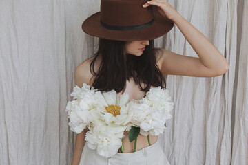 Stylish woman in hat posing with white peony flowers growing from pants. Floral fragrance. Self care