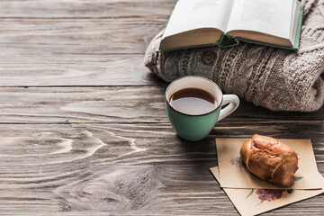 A cup of coffee on a wooden table, a warm sweater, an open book and a croissant. Lifestyle, cozy atmosphere. Copy space.