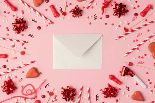 Top view photo of birthday composition red ribbon stars serpentine confetti heart-shaped cookies bright decorations and white closed envelope in the middle on pastel pink background with copyspace