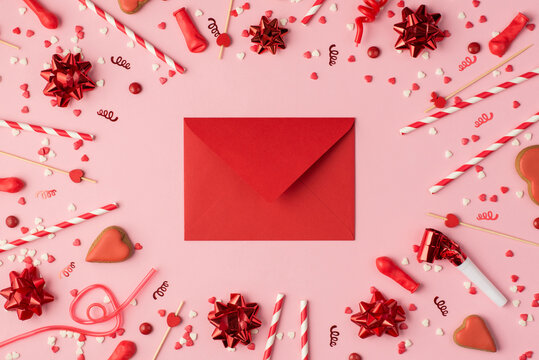 Top view photo of birthday composition with red ribbon stars serpentine confetti heart-shaped cookies bright decorations and red closed envelope in the middle on pastel pink background with copyspace