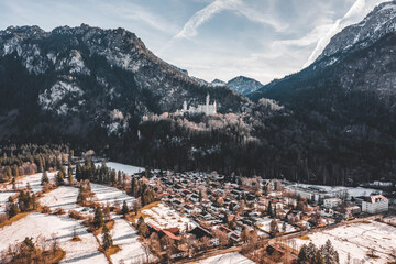 Aerial drone shot of Neuschwanstein Castle on snowy hill in winter sunlight in Germany with view of snowy village