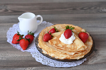 A white jug and pancakes with sour cream and strawberries stand on a wooden table on an openwork napkin. Image with selective focus.